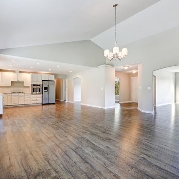Spacious rambler home interior with vaulted ceiling over glossy laminate floor. Empty light filled dining or living space adjacent to new white kitchen room features pale grey walls. Northwest, USA